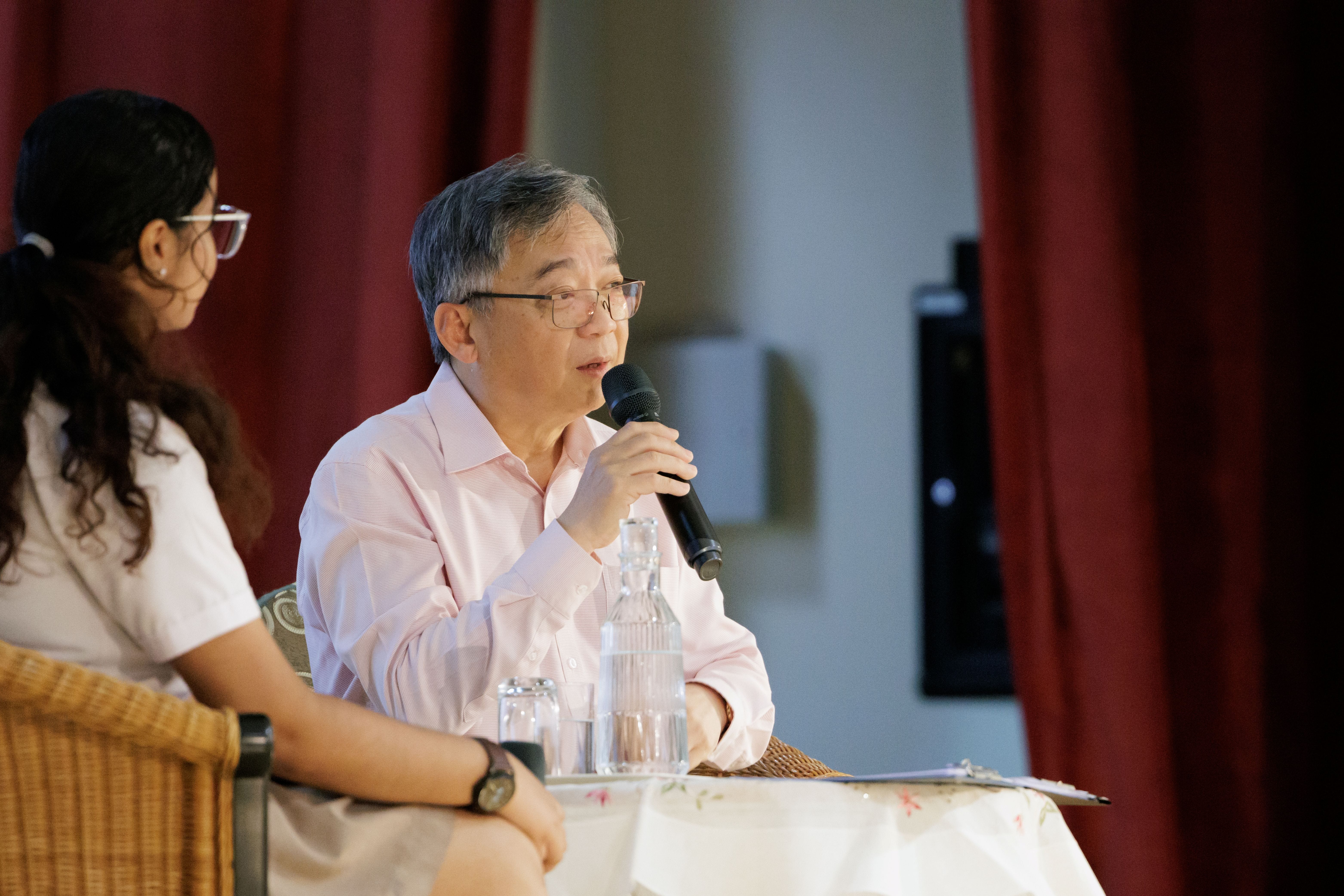 An older gentleman in a light pink shirt and glasses speaks into a handheld microphone while seated at a table with a glass water jug. A young female student sits beside him, listening attentively. Red curtains form the backdrop behind them.